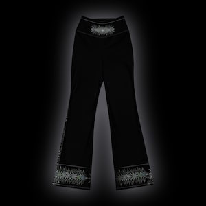May include: Black high-waisted pants featuring a wide-leg silhouette. The waistband and the lower hem of each leg are decorated with a detailed, symmetrical white and light green graphic pattern that resembles a stylized botanical design.