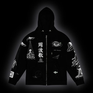 May include: A black zip-up hoodie featuring white graphic prints of flying saucers, extraterrestrial figures, a large human eye, and Japanese characters that read 周波数. The design includes a QR code on the sleeve and small geometric symbols.