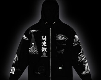 May include: A black zip-up hoodie featuring white graphic prints of flying saucers, extraterrestrial figures, a large human eye, and Japanese characters that read 周波数. The design includes a QR code on the sleeve and small geometric symbols.