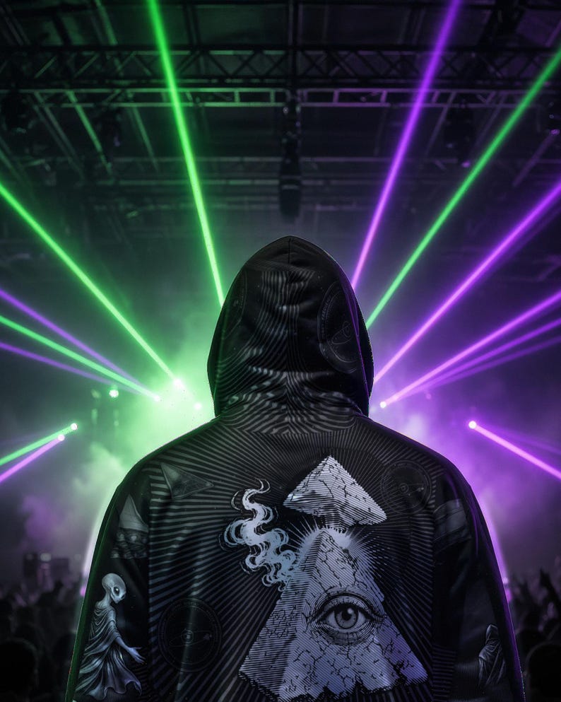 May include: A black hooded sweatshirt featuring a graphic print of an eye within a pyramid, a small alien figure, and abstract line patterns. The garment is shown from the back against a backdrop of bright green and purple laser light beams.
