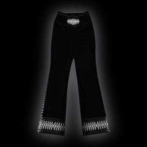 May include: Black high-waisted pants featuring a white geometric pattern at the waistband and a matching white decorative border along the bottom hem of each pant leg. The pants have a straight-leg cut and a solid black fabric construction.