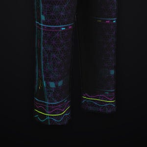 K&ouml;nnte beinhalten: Schwarze Hose mit einem sich wiederholenden violetten geometrischen Blume-des-Lebens-Muster. Das Design umfasst vertikale cyanfarbene Linien, horizontale neongelbe Wellenlinien sowie kleine rechteckige Akzente in Pink und Cyan.