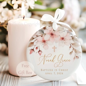 May include: A round white ceramic ornament featuring a delicate watercolor illustration of pink cherry blossoms and a small gold cross. The text reads Norah Grace, Baptized in Christ, and April 7, 2024, in elegant gold script and block lettering.
