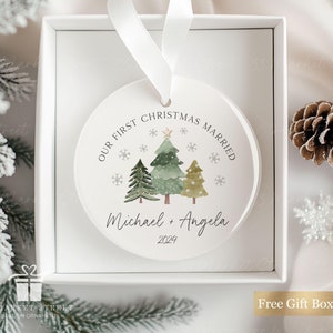 May include: White round Christmas bauble with a watercolour illustration of three green Christmas trees with gold stars. The text "OUR FIRST CHRISTMAS MARRIED" is written in a circle around the trees. Below the trees, the text "Michael + Angela 2024" is written.