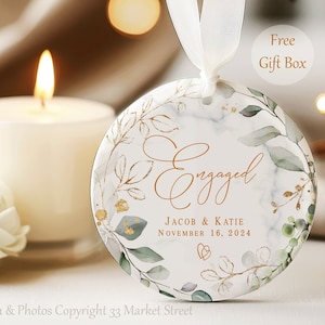 May include: Round, white, marble-patterned ornament with a gold leafy design and the text "Engaged" in gold script. The text "Jacob & Katie" and "November 16, 2024" are also in gold script.