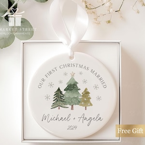 May include: A white ceramic Christmas bauble featuring a green watercolour tree design. The bauble reads "Our First Christmas Married" and "Michael + Angela 2024".