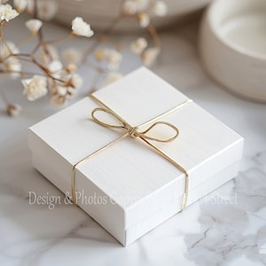 May include: A white gift box with a gold ribbon tied in a bow. The box is sitting on a white marble surface.