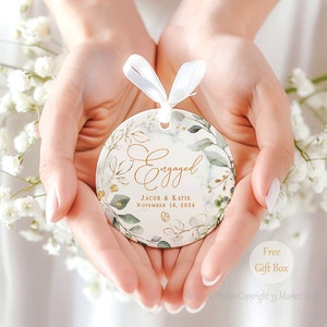 May include: White ceramic ornament with a gold and green floral design. The ornament says "Engaged" with the names "Jacob & Katie" and the date "November 16, 2024".