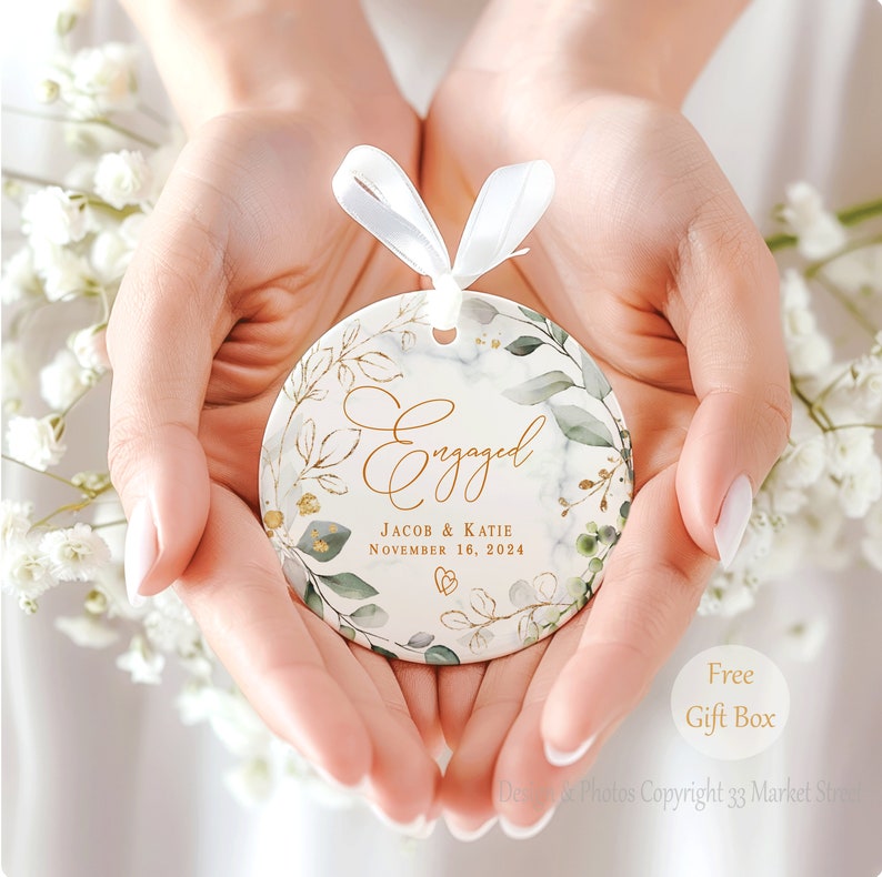 May include: White ceramic ornament with a gold and green floral design. The ornament says "Engaged" with the names "Jacob & Katie" and the date "November 16, 2024".