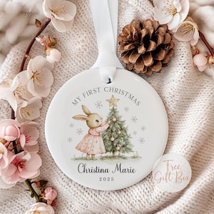May include: A round white ceramic ornament hanging from a white ribbon. The ornament features a graphic of a rabbit in a pink dress decorating a Christmas tree with a gold star. Text reads My First Christmas, Christina Marie, and 2025.