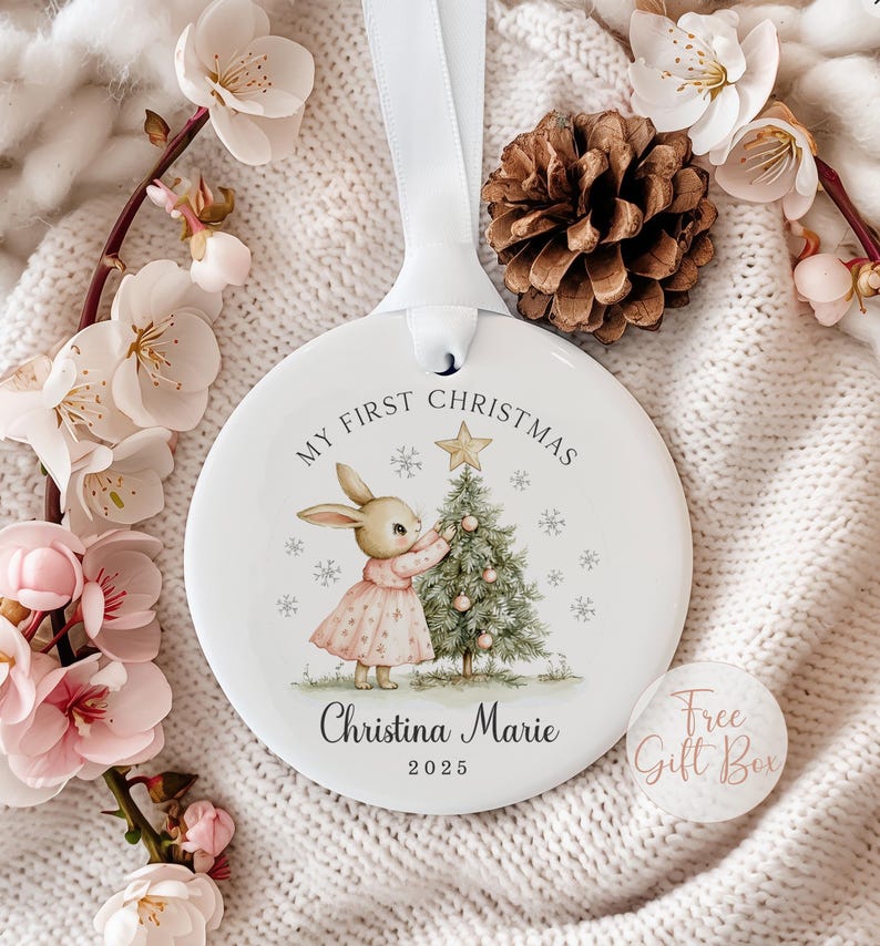 May include: A round white ceramic ornament hanging from a white ribbon. The ornament features a graphic of a rabbit in a pink dress decorating a Christmas tree with a gold star. Text reads My First Christmas, Christina Marie, and 2025.