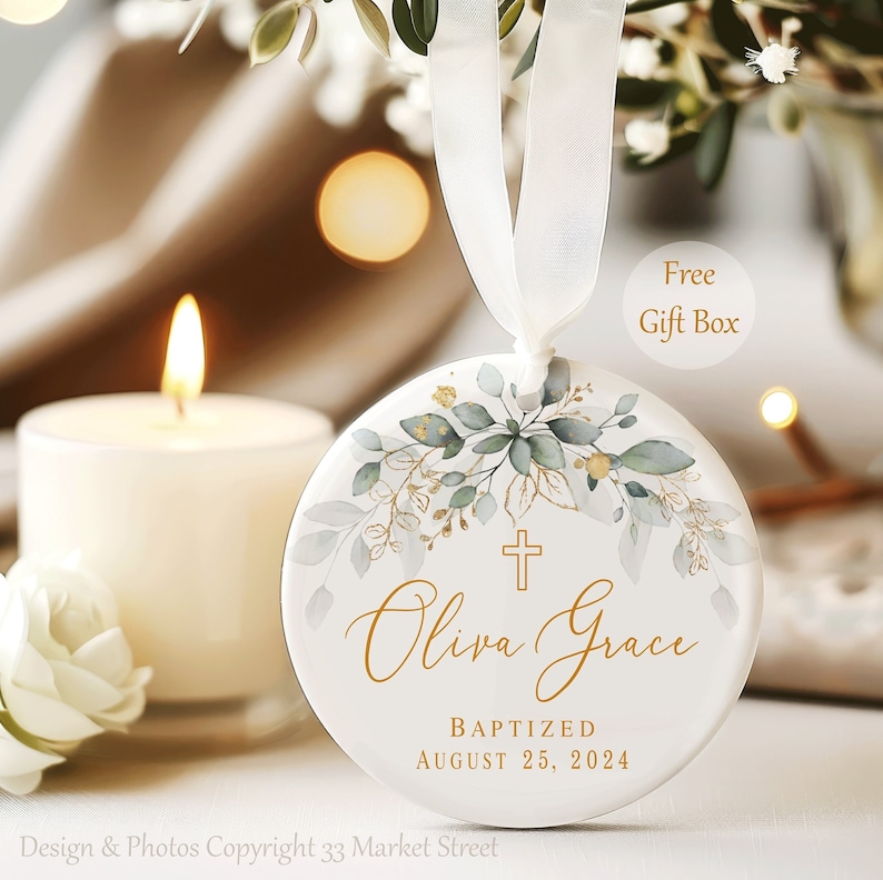 May include: White ceramic ornament with a gold cross, green and gold floral design, and the text "Oliva Grace Baptized August 25, 2024".