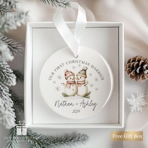May include: White round Christmas bauble with a watercolour illustration of two snowmen holding hands. The bauble says "Our First Christmas Married" and "Nathan + Ashley 2024".