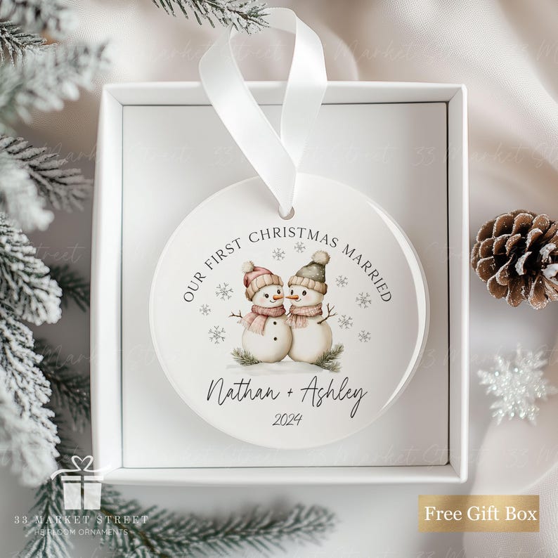 May include: White round Christmas bauble with a watercolour illustration of two snowmen holding hands. The bauble says "Our First Christmas Married" and "Nathan + Ashley 2024".