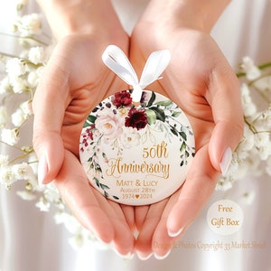 Op de afbeelding: Een rond wit keramieken ornament met een bloemmotief van bordeauxrode en zachtroze bloemen, groene bladeren en goudkleurige tekst 50th Anniversary, Matt & Lucy, August 28th, 1974 hart 2024, afgewerkt met een wit satijnen lint.