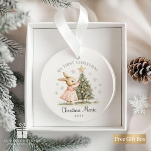 May include: A round white ceramic ornament with a white ribbon hanger, featuring a watercolor illustration of a rabbit decorating a Christmas tree. The text reads My First Christmas, Christina Marie, and 2024, surrounded by snowflakes.