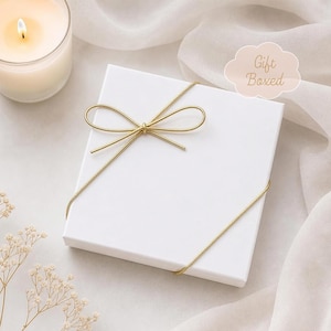Op de afbeelding: Een vierkante witte geschenkdoos vastgebonden met een dun goudkleurig koord in de vorm van een strik. De doos heeft een lichtroze wolk-vormige afbeelding met de tekst Gift Boxed in een schrijfletter op de bovenkant van het witte deksel.