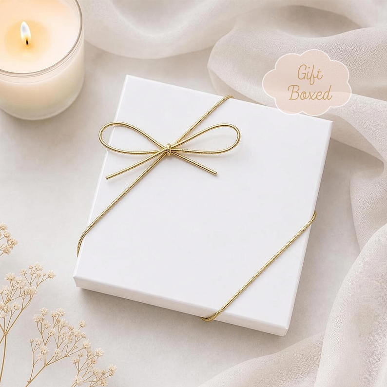 Op de afbeelding: Een vierkante witte geschenkdoos vastgebonden met een dun goudkleurig koord in de vorm van een strik. De doos heeft een lichtroze wolk-vormige afbeelding met de tekst Gift Boxed in een schrijfletter op de bovenkant van het witte deksel.