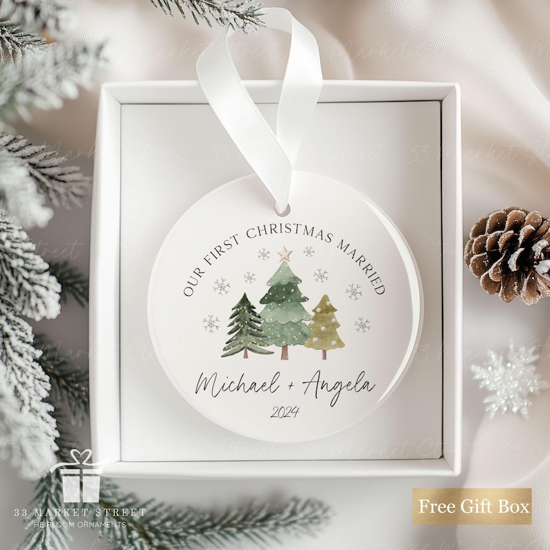 May include: White round Christmas bauble with a watercolour illustration of three green Christmas trees with gold stars. The text "OUR FIRST CHRISTMAS MARRIED" is written in a circle around the trees. Below the trees, the text "Michael + Angela 2024" is written.