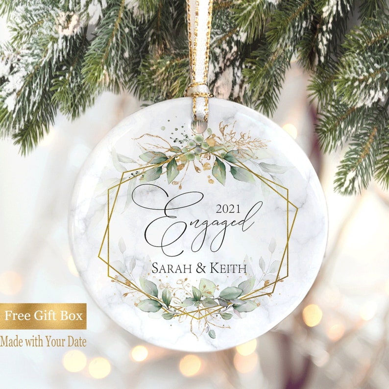 May include: White round ornament with a gold geometric frame and green foliage. The ornament says "2021 Engaged Sarah & Keith".
