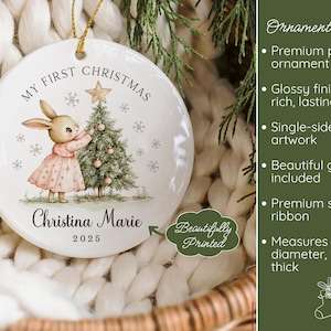 May include: A round white porcelain Christmas ornament measuring 3 inches in diameter and 1/8 inch thick. It features a bunny in a pink dress decorating a tree with the text My First Christmas, Christina Marie, and 2025 printed on the front.