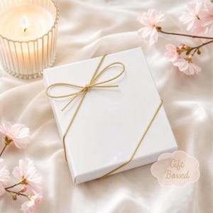 May include: A square white gift box tied with a thin gold cord in a bow, positioned next to a lit candle in a ribbed glass holder and light pink cherry blossom flowers. A small cloud-shaped tag with the text Gift Boxed is visible.