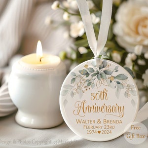 Op de afbeelding: Een rond wit keramieken ornament met gouden tekst die 50th Anniversary, Walter & Brenda, February 23rd, 1974 hart 2024 leest. Het ontwerp bevat groene aquarelbladeren en gouden accenten, opgehangen aan een wit satijnen lint.