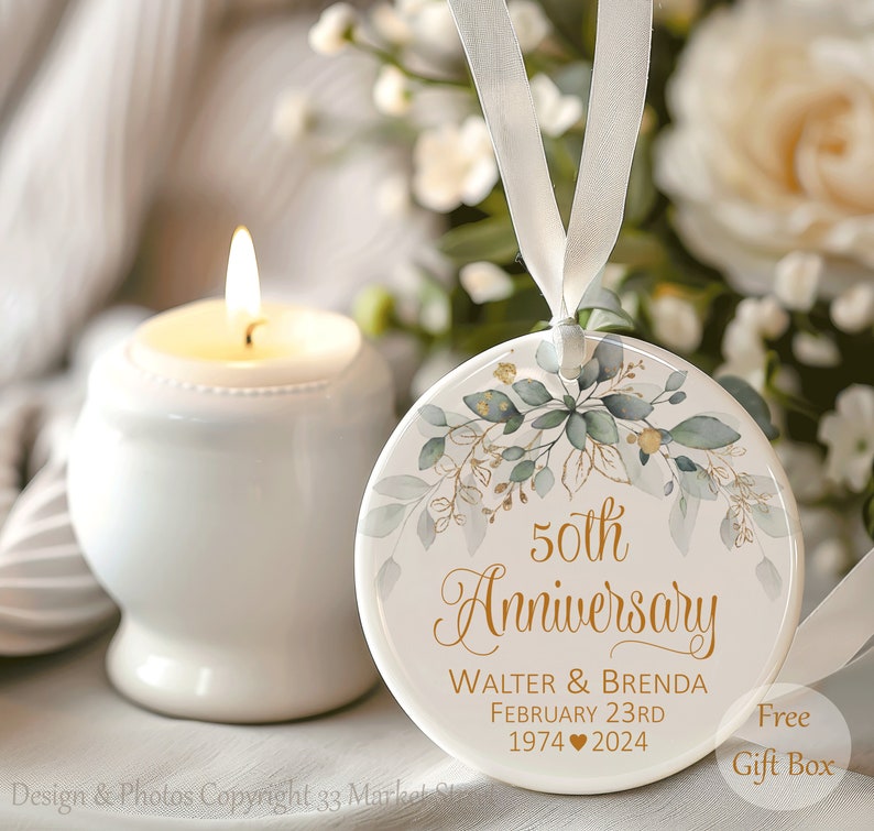 Op de afbeelding: Een rond wit keramieken ornament met gouden tekst die 50th Anniversary, Walter & Brenda, February 23rd, 1974 hart 2024 leest. Het ontwerp bevat groene aquarelbladeren en gouden accenten, opgehangen aan een wit satijnen lint.
