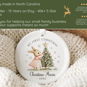 May include: A round white ceramic ornament featuring a printed illustration of a rabbit in a pink dress decorating a Christmas tree. The text reads My First Christmas, Christina Marie, and 2025. The ornament has a small hole at the top for hanging.