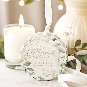 May include: A round, white ceramic ornament with a gold inscription that reads "Engaged" with a leafy green and gold watercolor design. The text below the inscription reads "Jacob & Katie November 16, 2024".