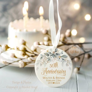 Op de afbeelding: Een rond wit keramieken ornament hangend aan een wit lint. Het ornament is voorzien van een botanisch aquarelontwerp in groen en goud, met de tekst 50th Anniversary, Walter & Brenda, February 23rd, 1974 en 2024 in gouden letters gedrukt.