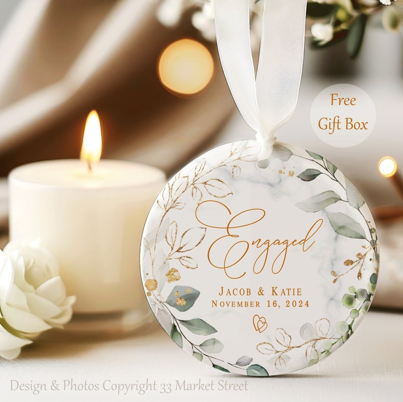 May include: Round, white, marble-patterned ornament with a gold leafy design and the text "Engaged" in gold script. The text "Jacob & Katie" and "November 16, 2024" are also in gold script.