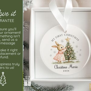 May include: A round white ceramic Christmas ornament featuring a printed illustration of a rabbit in a pink dress decorating a Christmas tree. The text reads My First Christmas, the name Christina Marie, and the year 2024. It hangs by a white ribbon.