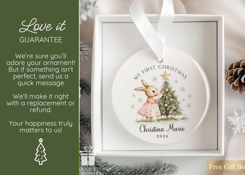 May include: A round white ceramic Christmas ornament featuring a printed illustration of a rabbit in a pink dress decorating a Christmas tree. The text reads My First Christmas, the name Christina Marie, and the year 2024. It hangs by a white ribbon.