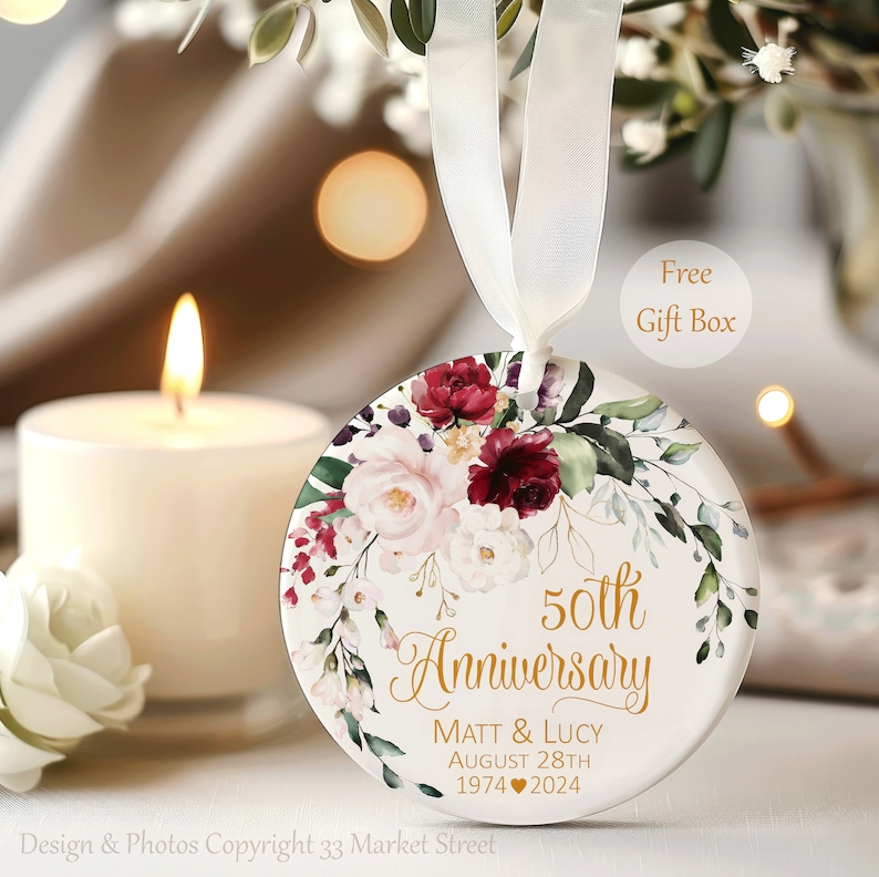 Op de afbeelding: Een rond wit keramieken ornament hangend aan een wit lint, voorzien van een bloemstuk van bordeauxrode en zachtroze rozen. De tekst luidt 50th Anniversary, Matt & Lucy, August 28th, 1974 hart 2024 in gouden script en blokletters.