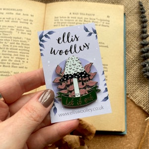 May include: A green and white enamel pin featuring a mushroom with dripping black liquid and pink leaves. The pin has a green banner with the text "Let it out" and the website address "www.elliswoolley.co.uk".