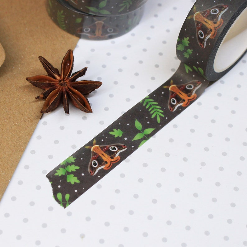 May include: A roll of black washi tape with a repeating pattern of brown moths and green leaves. The tape is on a white background with small gray dots.