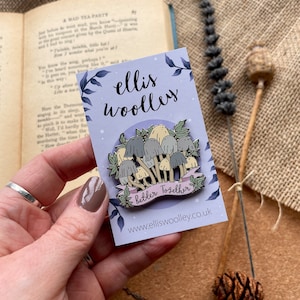May include: A silver enamel pin featuring a cluster of mushrooms with a banner that reads "Better Together". The pin is on a purple background with the text "ellis woolley" and a website address.