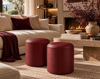 Peut inclure: Deux poufs cylindriques recouverts de cuir rouge profond avec une surface supérieure lisse et arrondie. Ces tabourets présentent un design solide et structuré ainsi qu'une couleur cramoisie riche et uniforme, posés sur un tapis beige texturé.