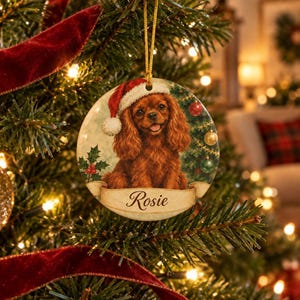 Op de afbeelding: Een ronde keramische kerstornament met een bruine spaniël die een rood-witte kerstmuts draagt. De hond staat tegen een feestelijke achtergrond met hulst en kerstballen. Een banner onderaan toont de naam Rosie in een sierlijk lettertype.