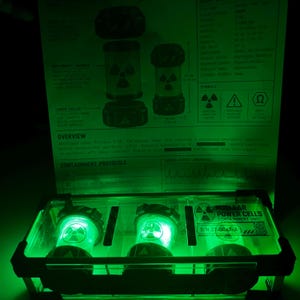 May include: A clear rectangular containment unit holding three glowing green cylindrical nuclear power cells. The unit features black corner brackets and a label reading NUCLEAR POWER CELLS CONTAINMENT UNIT S/N 27-0047-A with a QR code.