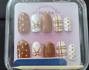 May include: A set of ten press-on nails in a clear plastic case labeled Collection Nail Art. The nails feature brown and white colors with polka dot, bow, and plaid patterns, including small rhinestones. A blue nail file and wooden stick are included.