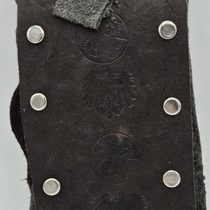 May include: Close-up of a dark brown leather item with silver metal button closures. The leather has embossed designs, possibly a crest or emblem. A gray fabric detail is visible.