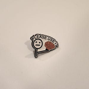May include: A white enamel pin featuring a black-outlined stick figure with a neutral expression, holding a pink brain illustration. The text PLEASE USE IT is printed in black capital letters along the top edge of the pin design.
