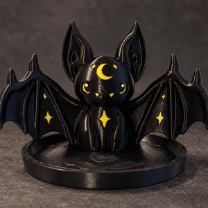 May include: A black bat figurine with large ears and outstretched wings, featuring yellow accents including a crescent moon on its forehead, small fangs, and four-pointed star shapes on its chest and wings, set on a circular black base.