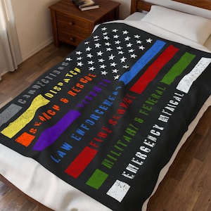 May include: A black fleece blanket featuring a stylized American flag design with white stars and colored stripes. Text includes CORRECTIONS, DISPATCH, SEARCH & RESCUE, SECURITY, LAW ENFORCEMENT, FIRE SERVICE, MILITARY & FEDERAL, and EMERGENCY MEDICAL.