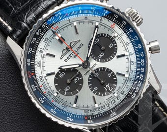 Puede incluir: Un reloj cronógrafo Breitling Navitimer con esfera plateada efecto rayos de sol, tres subesferas negras y un bisel azul con regla de cálculo. El reloj cuenta con una caja de acero inoxidable y una correa de cuero negro con costuras blancas.