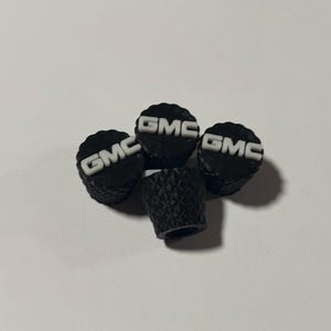 May include: A set of four black tire valve stem caps with a textured, diamond-patterned side surface. Each cap features a white, raised GMC logo on the flat top. The caps are cylindrical in shape and designed for automotive tire valves.
