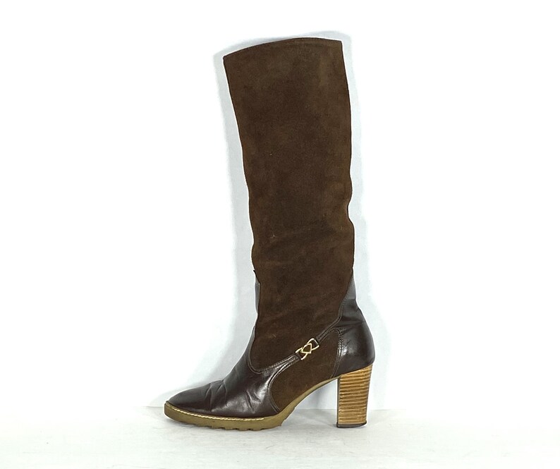 May include: Brown suede knee-high boots with a block heel and a buckle detail on the side.