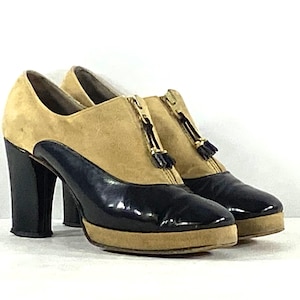 May include: A pair of two-tone platform shoes featuring a tan suede upper and a black patent leather lower section. The shoes have a front zipper closure with a small black tassel pull, a thick block heel, and a tan platform sole.